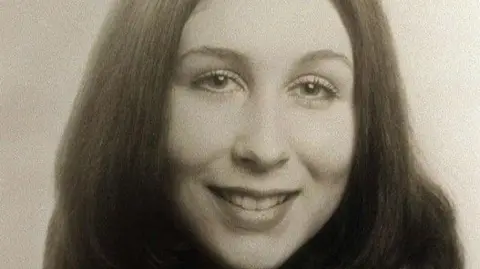 Black and white photo of Debra-Anne Cartwright. She has shoulder length dark hair parted in the middle. She is smiling. 