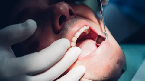 Getty Images Close-up of a man having his teeth examined by a dentist who is wearing white latex gloves and looking in his mouth using a dental mirror.