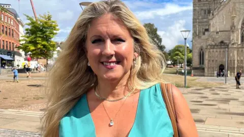 BBC Alison Sheridan, a Conservative member of Exeter City Council, wearing a turquoise dress, a silver necklace and a second necklace with a jewel and the brown straps of a handbag over her shoulder with the front of Exeter Cathedral in the background.