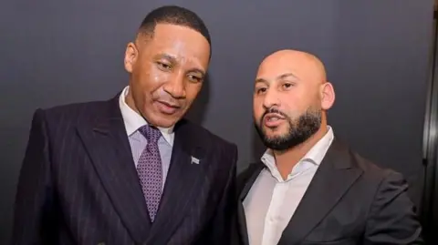 Demetrios Bradshaw Bradshaw is wearing a dark suit with an open-collared white shirt. He is standing next to a man wearing a lilac shirt, purple tie and black suit jacket.