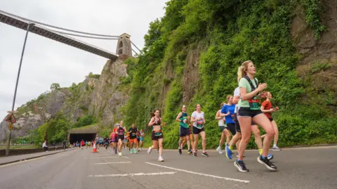 AJ Bell Great Bristol Run A group of athletes running down the Portway in Bristol. The Clifton Suspension Bridge can be seen in the background.