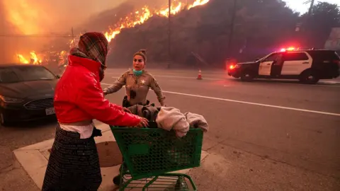 A police officer escorts a homeless woman pushing her belongings on a trolley, on a street with a police car and burning mountain in the background, in Topanga Canyon Blvd.