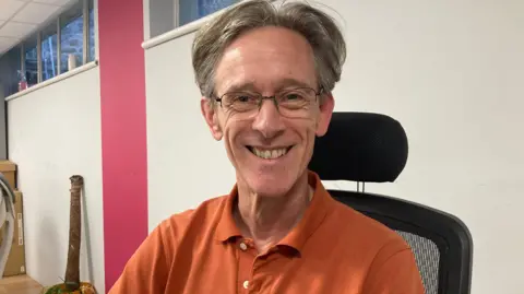 BBC A mid shot of Marko Kennedy. He has short grey hair and is wearing glasses, and an orange polo shirt.