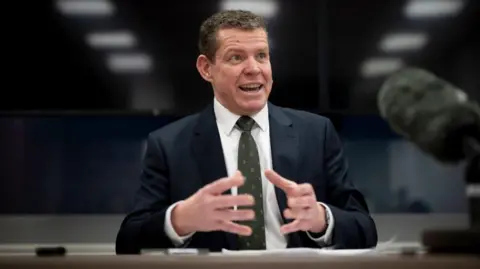 Rhun ap Iorwerth, sat in a press conference on Tuesday morning, gesticulating with his hands while sat at a desk in front of a boom microphone.