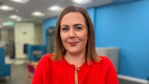 Rachel Spiers. She has shoulder length light brown hair and brown eyes, wearing a red buttoned cardigan.