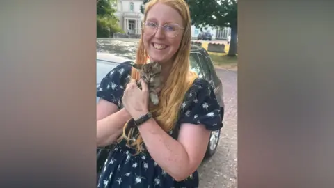 Devon and Somerset Fire Service Amy Down has sandy red hair and is wearing a navy dress with white flowers on it. She is holding a very small dark and light brown tabby kitten which has just been rescued.