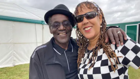 A man and woman stand in a field with their arms around each other. The man has short curly grey hair and he is wearing a black polo shirt and a black pork pie hat. The woman has curly brown hair and is wearing a black and white checked jumper. 