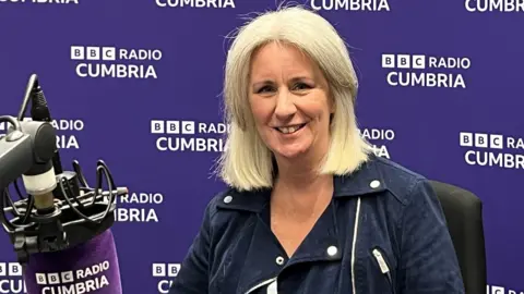 Heather Wainwright sitting in a BBC Radio Cumbria studio, with a branded purple microphone in front of her and a branded purple wall behind her. She has blonde/white hair in a bob and is wearing a blue jacket. She is smiling at the camera.