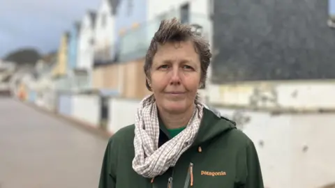 A woman wearing a green Patagonia rain jacket standing outdoors, near a row of properties. The colourful properties are blurred out. 