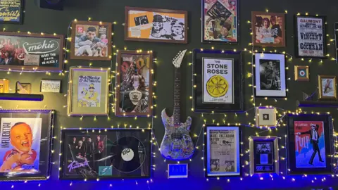 Grace Wood/BBC A green pub wall covered in posters from gigs in Bradford, an electric guitar and fairy lights