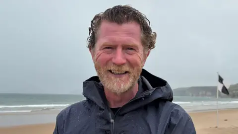 Andrew Todd stands on Whiterocks beach on Portrush. He has wavy brown hair and a ginger/grey beard. He is wearing a dark blue raincoat. It is an overcast day.