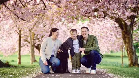 Bethany Clarke A man, woman and their young son pose for a photo with their black Labrador under the blossom trees