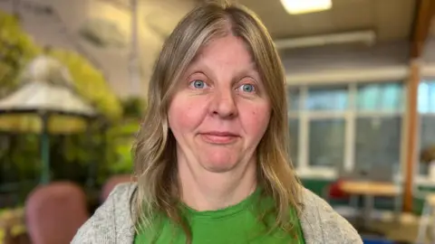 Caroline Hickman is standing in the Endike Community Care centre. She is staring ahead and she has shoulder length blonde her with a green t-shirt and a grey cardigan. Behind her are empty chairs inside the daycare centre which has now been forced to close.