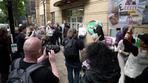 A group of people stood outside Notting Hill Genesis HQ, looking at a man holding a microphone, speaking to the crowd, and one woman on the left side is holding up a large placard.