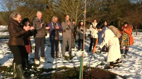 A woman with a spade planting a tree surrounded by a group of people who are clapping. There is snow on the ground and trees behind, some are bare and some have leaves. 