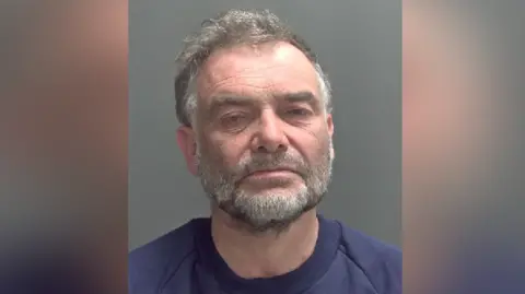 Norfolk Police Image shows mugshot of man in purple jumper, he has a grey beard