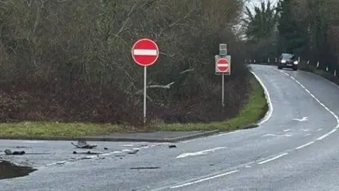 A dual carraigeway is surrounded by trees. A no entry sign can be seen. A car with its lights on is in the distance. 