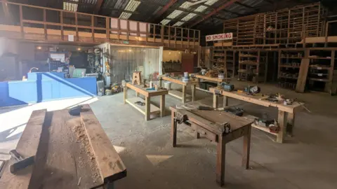 Ben McConnell A set of workbenches are in the middle of a warehouse. Hammers and woodchips can be seen on them. A large sign on the far side of the room reads "no smoking".