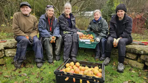 Aisha Iqbal/BBC Five people of mixed ages, wearing outdoor clothing, sit in a semi circle on a low dry stone wall. There are gardening tools around them. Two women in the centre are holding a crate of apples, and another crate of apples is on the grass in front of the group.