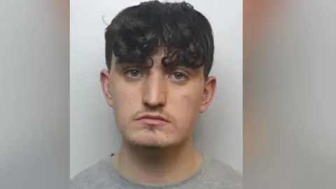 Mugshot of Ethan Baily. He has short dark curly hair and a small amount of stubble on his upper lip and chin. He is wearing a grey t-shirt and is looking at the camera with a blank expression.