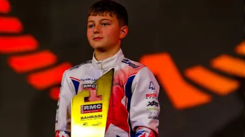 Racing Numbers A young boy dressed in white racing overalls standing on a podium holding a gold-coloured trophy, with the number one on it. He is looking out towards the crowd and smiling.