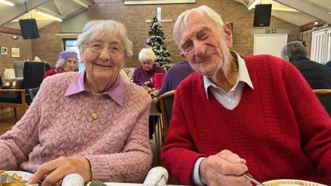 Two people looking into the camera smiling, as they hold their knives and forks in front of their Christmas dinner, as if the photo is taken before they take a bite. They are leaning towards each other affectionately. The woman, on the left, is wearing a pale pink knitted jumper with a darker pink shirt underneath. The man on the right, is wearing a red knitted v neck jumper. A white and dark blue striped shirt is underneath. They're in a community centre hall and behind them is a long table with diners and in the far background, a Christmas tree and a big silver cross on the wall.