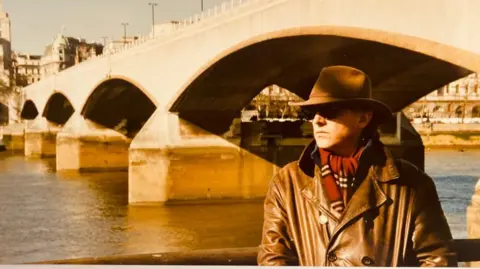 Bob Baldwin Rik Mayall, not easily identifiable as he is wearing a fedora-style hat and sunglasses, standing wearing a brown leather coat and a red scarf. He is standing by a river with a large bridge over it.