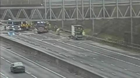 National Highways Several lanes of the M25 motorway, showing a van, people standing around it, a car on the other side, fire engines and an overhead metal gantry. 