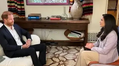 Prince Harry, right, sits on a white sofa in a living room opposite Nada Tawfik, left, who sits on a striped orange couch.