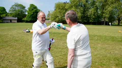 PA Media Ed Davey fist bumps Ian Thorn (who we can only see the back of) while they are both in cricket kit. They are on a cricket field with trees in the background on a sunny day.