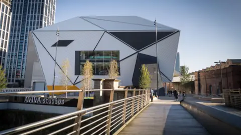 Aviva Studios, a grey building made of triangular panels. A bridge runs over a canal towards the site while people can be seen milling around a forecourt. 