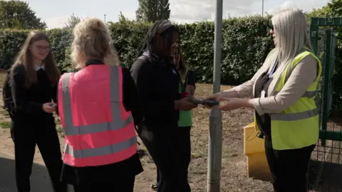 The photo shows two school pupils approaching some green school gates. Greeting them are two women, both in hi-vis jackets. Their arms are reaching out to touch black pouches. 