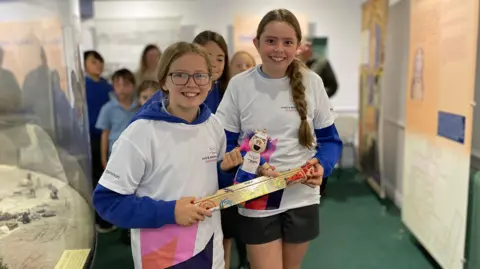 Two girls wearing white t-shirts over blue jumpers are together holding the baton and a soft toy mascot. They are standing in a museum exhibition space and there are more children walking behind them.