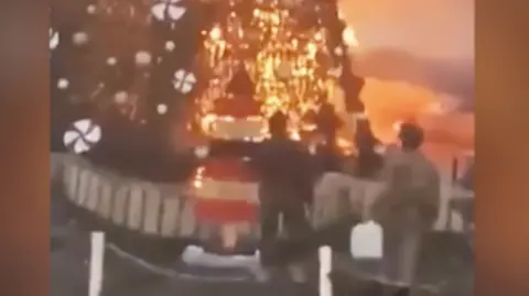 In a still from a video, two men stand at the base of an enormous christmas tree, which is on fire