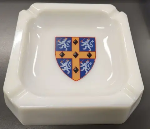 The Story/Durham County Council A square white ashtray with the County Durham logo in the centre. The logo is formed from a blue shield bearing a gold cross and decorated with five black diamonds. In each of the four blue sections, there is a prancing lion wearing a crown and carrying a sword