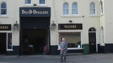 Nicholson family A man in a grey suit standing in the street in front of a cream and black building. The sign on the building reads DAVID DUGGLEBY VALUERS.