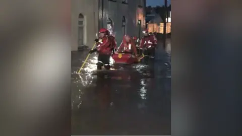 Antrim Guardian A nighttime photo showing a dinghy in the middle distance with a man in it wearing an orange coat and buoyancy aid. The dinghy is red. It is being pushed through floodwater in a residential street flanked by people in red life jackets and red helmets.
