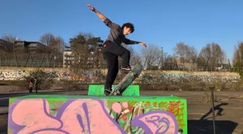 A boy skates on a graffiti-covered green mini-ramp in a skatepark, set against a blue sky.