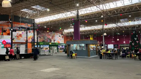 Grace Wood/BBC A cluster of food stalls in an indoor market hall. Chairs and tables can be seen in the distance, while a performance stage is to the left.