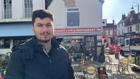 Tom Jackson/BBC Hussain Aslan is standing in front of the Market Cafe, which has red signage and outdoor seating. He is smiling at the camera while wearing a black puffer coat. He has black hair and facial hair.
