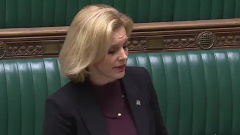 UK PARLIAMENT Woman infront of Parliament's green benches with short blonde hair purple top black blazer 