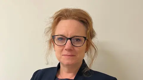 Suffolk County Council Sarah-Jane Smedmor smiles at the camera. She has red hair that has been tied back with some loose bits framing her face. She wears glasses and a navy jacket. 