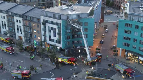 James Robert An aerial photo showing eight fire engines stopped in a town square, with a block of flats on the corner blackened by fire. There's smoke still rising from the building, with a ladder rising from one of the fire engines.