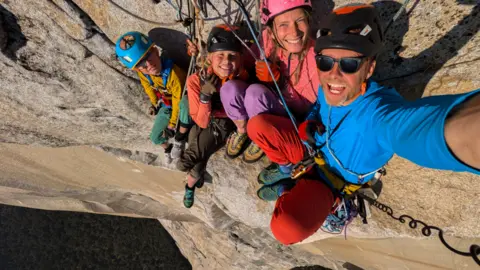 Leo Houlding Leo Houlding and his family on the side of El Capitan. The family of four are wearing brightly coloured climbing attire in bright blues, reds, pinks, purples and greens, and have on outdoor gear which include helmets. They are sort of sitting on the side of the huge, cream-coloured granite rock face which is really exposed and are taking a selfie, which means our view of them is from above, making the huge drop below them even more evident.