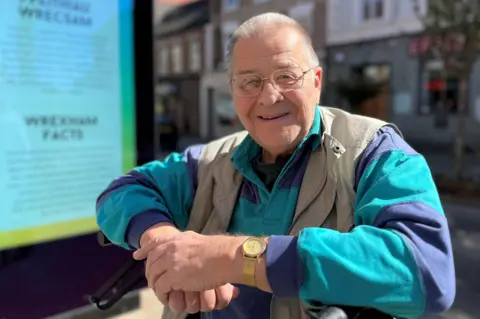 Ronald is in his 70s with a grey buzz cut wearing a blue and teal rugby shirt with a khaki gilet over. He is wearing gold framed glasses and a gold watch and is smiling. He is sitting on a walking frame, just visible, next to an information board which reads Wrexham Facts and has some out of focus text. 