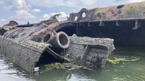 A rusty green, gray and red wreck of a German submarine lies in green water.