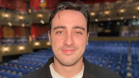 Helen Mulroy/BBC A man sat in front of tiers of blue theatre seating