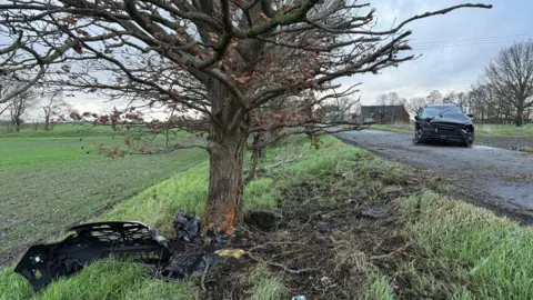 CAMBRIDGESHIRE POLICE Part of the front bonnet of the black car is lying in front of a tree with largely bare branches. A small amount of bark from the tree has been damaged. There is a trail of mud leading from the tree across a grass verge to a road where the car is sitting. It has a crunched up bonnet. 
