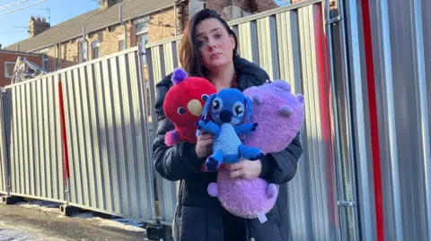 BBC Grace Porter holding stuffed animal toys outside the blast damaged home. She has long, straight brown hair and is wearing a black puffer jacket. One of the houses behind her has its roof and upper floor blown off. The site is fenced-off. 