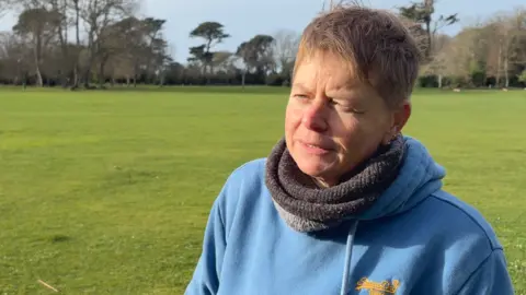 Sarah is in a park with green grass and trees in the background. She is wearing a blue hoodie and has short light brown hair.
